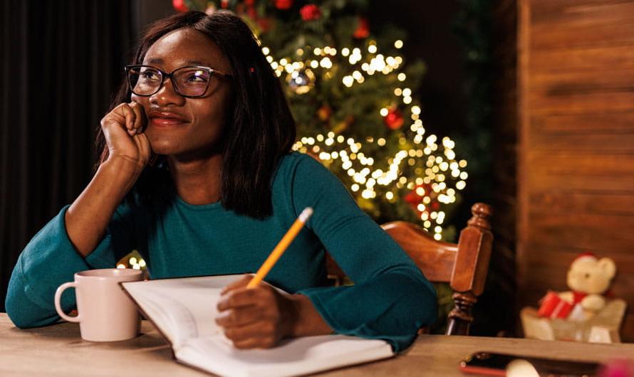快乐的年轻女子坐在家里的办公桌旁, 思考她的新年决心，并把它们写在日记里.